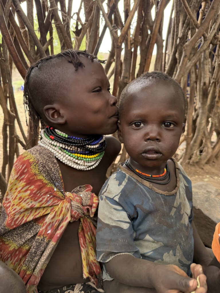Das Foto zeigt ein Mädchen aus Nakolonyoi die vermutlich ihrem Geschwister ein Küsschen auf die Wange gibt (Busu busu). Die Nahaufnahme fängt einen zärtlichen Moment zwischen zwei kleinen Kindern ein, die eng beieinandersitzen. Beide haben ernste, aber unschuldige Ausdrücke, mit großen Augen und leichten Schmutzspuren auf der Haut. Die Szene vermittelt Themen wie familiäre Zuneigung, Kindheit in Armut und kulturelle Elemente in einer ruralen, afrikanischen Dorfgemeinschaft wie Nakolonyoi, mit einem Fokus auf Emotion und Nähe in einer einfachen Umgebung.