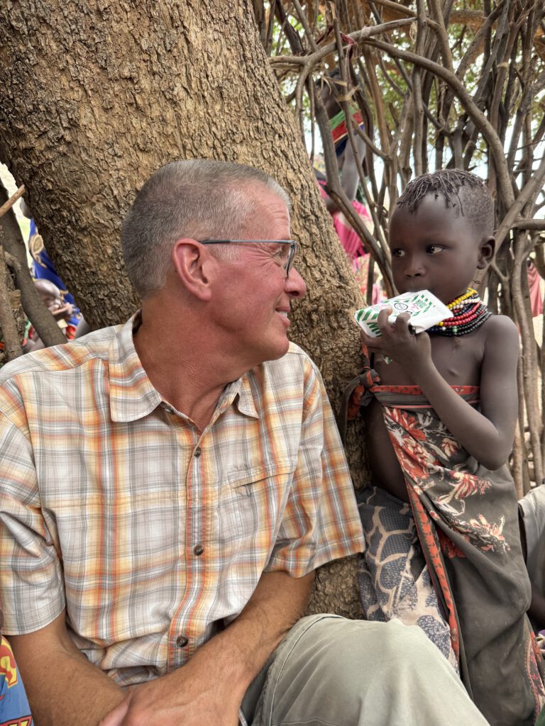 Achim sitzt neben einem kleinen Mädchen aus Nakolonyoi, das eine Tüte Milch in der Hand hält. Die beiden schauen sich tief in die Augen und lächeln einander an. Die Szene strahlt Nähe und Vertrauen aus.