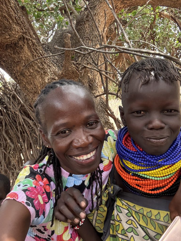 Patricia und eine junge Frau aus Nakolonyoi lächeln in die Kamera. Die junge Frau trägt traditionellen bunten Halsschmuck. Beide wirken fröhlich und halten sich eng umarmt.