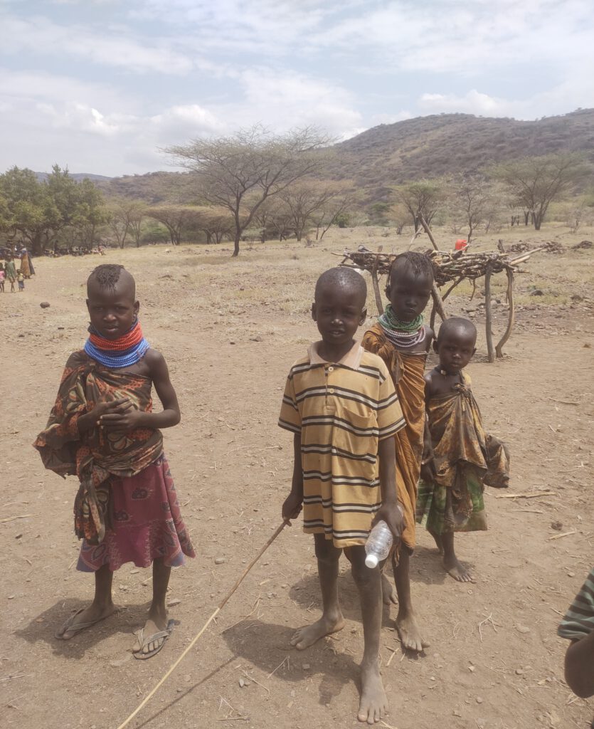 Das Foto zeigt kids aus Nakolonyoi die auf Essen und Trinkwasser hoffen. Die Kinder tragen einfache Kleidung, einige halten Stöcke oder kleine Gegenstände in den Händen und sind barfuß. Ihre Ausdrücke wirken ernst und erwartungsvoll, während sie direkt in die Kamera blicken. Die Szene ist geprägt von Trockenheit und Bedarf an humanitärer Hilfe und unterstreicht Themen wie Armut, Wassermangel und die Hoffnung der lokalen Gemeinschaft auf Unterstützung.