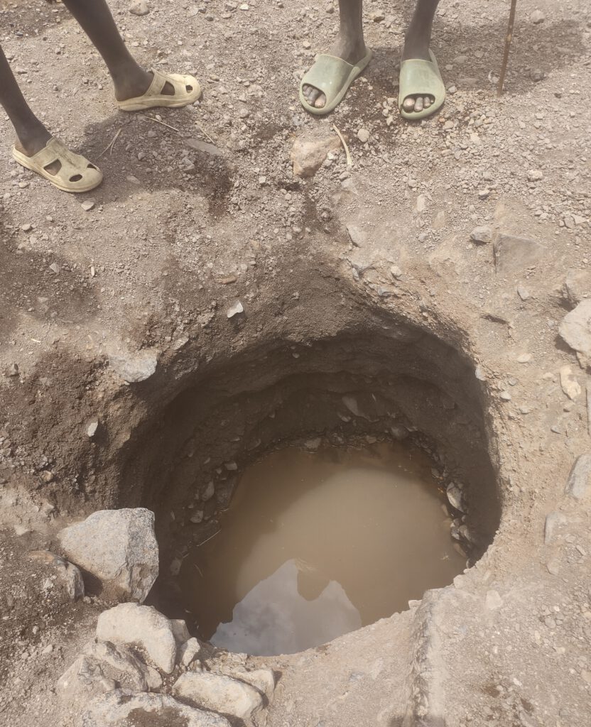 Das Foto zeigt die kläglichen Wasserreste in einem Loch im Dorf Nakolonyoi. Im Vordergrund sind zwei Paar Beine und Füße in einfachen Flip-Flops zu sehen, die am oberen Rand eines etwa meterbreiten, handgegrabenen Lochs im staubigen Boden stehen. Das Loch führt senkrecht nach unten und enthält am Grund eine kleine Pfütze trüben, schlammigen Wassers, umgeben von lockeren Erdbrocken und Steinen. Die Szene vermittelt Trockenheit und Armut in der ruralen, Turkana Region.