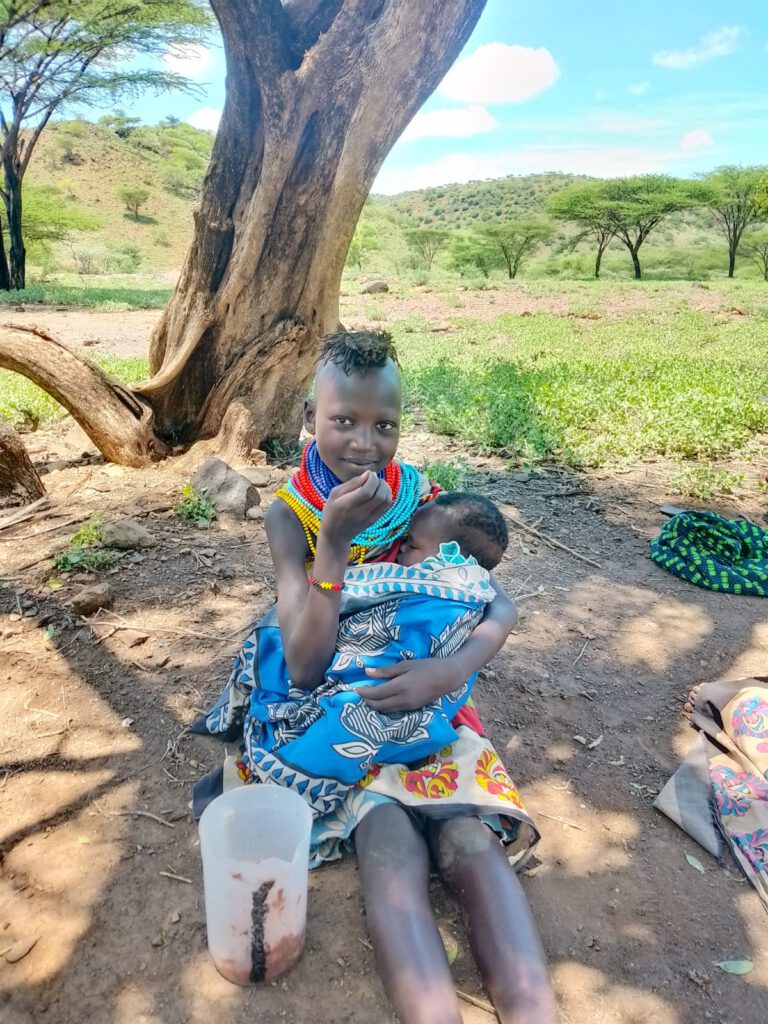 Das Foto zeigt eine junge Mutter aus Nakolonyoi glücklich und dankbar für die Lebensmittel die wir gebracht haben. Sie sitzt entspannt auf dem erdigen Boden im Schatten eines großen, knorrigen Baums. Sie hält ein Baby liebevoll im Arm, das in ein blaues Tuch gewickelt ist und friedlich schläft. Mit der rechten Hand führt sie etwas Mais-Bohnenbrei zum Mund. Die Szene strahlt Wärme und Zufriedenheit aus, unterstreicht Themen wie mütterliche Fürsorge, Gemeinschaftshilfe und den positiven Impact humanitärer Lieferungen in einer ruralen, afrikanischen Region.