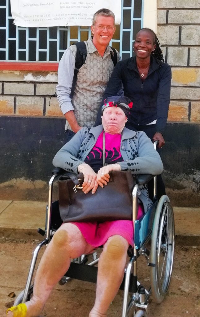 Das Foto zeigt Achim und Patricia von BlackAndWhite und die Albino Dame Josphin. Im Vordergrund sitzt Josphin, eine Frau mit Albinismus, in einem nagelneuen Rollstuhl auf einem staubigen Boden; sie hat helle Haut, ist barfuß mit gelben Schuhen, und mit staubigen- schmutzigen Knien. Hinter ihr steht links Achim der freundlich lächelt und rechts daneben Patricia die ebenfalls lächelt Die Szene strahlt Wärme, Unterstützung und Inklusion aus, mit natürlicher Sonnenbeleuchtung und Themen wie Behinderung, Albinismus und Gemeinschaftshilfe in einer armen, ruralen Umgebung.