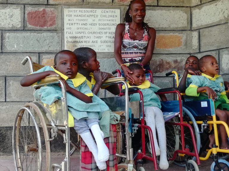 Das Foto zeigt Patricia von BlackAndWhite mit Kindern der Daisy Special School, Kakamega. Im Vordergrund sitzen fünf Kinder mit körperlichen Behinderungen in einer Reihe von Rollstühlen auf einem betonierten Boden; sie tragen einheitliche hellblaue Shirts mit gelben Kragen, kurze Hosen oder Röcke und Socken, und blicken ernst oder neugierig in die Kamera. Die Kinder wirken jung, etwa 5-10 Jahre alt, einige lehnen sich an oder halten sich an den Armlehnen fest. Hinter ihnen steht Patricia, die freundlich lächelt und eine Hand auf die Schulter eines Kindes legt. Die Szene unterstreicht Themen wie Inklusion, Bildung für behinderte Kinder und humanitäre Arbeit in einer kenianischen Region, mit Fokus auf Gemeinschaft und Fürsorge.