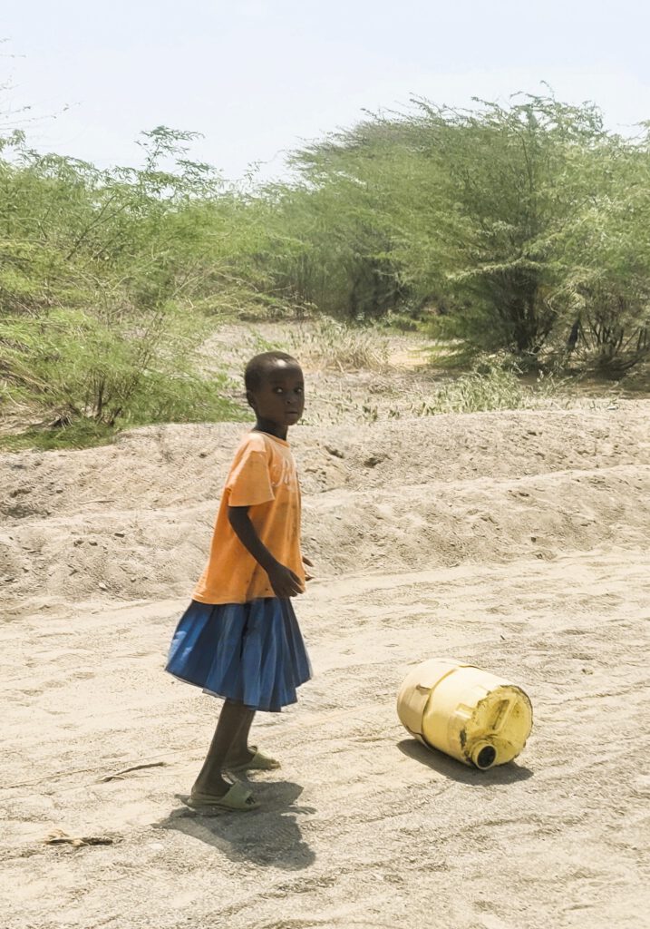 Das Foto zeigt ein kleines Mädchen welches mit Anstrengung eine große, gelbe 20 L Plastiktonne auf dem staubigen, unebenen Boden rollt um Wasser zu holen. Die Szene vermittelt Themen wie Kindheit in Armut, tägliche Herausforderungen in ländlichen Gebieten Afrikas und die Notwendigkeit, weite Wege für grundlegende Ressourcen zurückzulegen und betont die Isolation und Härte des Alltags.