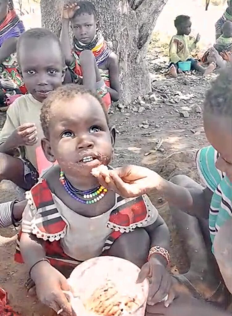 Das Foto zeigt ein kleines Mädchen etwa 2-3 Jahre alt, welches dankbar einen Brei aus Mais und Bohnen isst und nun noch um etwas Wasser bittet. Sie blickt mit großen, ausdrucksstarken Augen leicht nach oben, was eine Bitte oder Dankbarkeit vermittelt. Umgeben ist es von anderen Kindern, die ebenfalls sitzen, einige mit traditionellen Halsringen und einfacher Kleidung, unter den schattigen Ästen eines Baums im Dorf Nakolonyoi. Die Szene unterstreicht Themen wie Hunger, humanitäre Hilfe und den täglichen Kampf um Nahrung und Wasser in armen, ruralen Regionen Afrikas, mit einer warmen, natürlichen Beleuchtung und Fokus auf die Emotionen der Kinder.