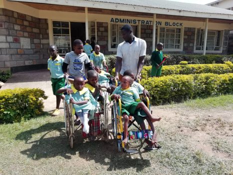 Kinder und Betreuer der Daisy Special School stehen vor dem Schulgebäude. Einige Kinder sitzen in den neu gespendeten Rollstühlen, während Betreuer und andere Kinder sie begleiten.
