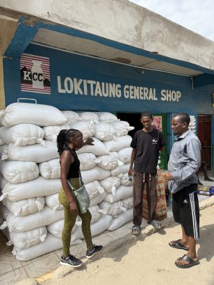 Das Foto zeigt Patricia beim Großhändler in Lodwar wo wir die 11 Tonnen Lebensmittel gekauft haben. Im Vordergrund steht Patricia die energisch gestikuliert und mit zwei Männern spricht – dem Großhändler und Mitarbeiter, sie stehen entspannt da und hören zu. Links von ihnen ist eine hohe Wand aus gestapelten weißen Säcken zu sehen, die mit Mais und Bohnen gefüllt sind, Der Laden "LOKITAUNG GENERAL SHOP" hat eine blaue Fassade mit einem roten Logo und ist in einer staubigen, urban-ruralen Straße in Lodwar angesiedelt Die Szene unterstreicht die Verhandlungsituation, im Kontext humanitärer Hilfe für Dörfer wie Nakolonyoi. Die Atmosphäre wirkt geschäftig und kooperativ