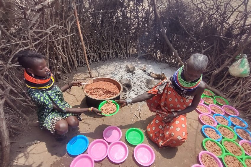 Das Foto zeigt 2 Frauen aus dem Dorf Nakolonyoi in ihrer einfachen Manyatta Küche, wie sie die Teller die BlackAndWhite gekauft hatten mit Mais und Bohnen befüllen. Die Szene spielt in einer traditionellen, runden Hütte aus verflochtenen Zweigen und Ästen, mit einer zentralen Feuerstelle, auf der Asche und ein schwarzer Topf mit dem Brei aus gelblichen Maiskörnern und Bohnen stehen. Links kniet eine junge Frau in einem grün-blau gemusterten Kleid mit farbigen Perlenhalsringen in Rot, Gelb und Blau, die einen pinken Plastikteller hält und portionsweise Essen schöpft. Rechts sitzt eine ältere Frau in einem orange-weißen gepunkteten Tuch mit ähnlichen Halsringen, die einen Löffel in der Hand hält und den Topf rührt, während sie den gefüllten Teller anreicht. Vor ihnen liegen zahlreiche bunte Plastikteller in Pink, Blau, Grün und Gelb auf dem staubigen Boden, einige bereits mit Essen gefüllt, was auf eine Verteilung an die Gemeinschaft hinweist. Die Atmosphäre ist rustikal und authentisch, mit natürlicher Beleuchtung durch Lücken in den Wänden, die die Härte des Alltags in einer arid afrikanischen Region unterstreicht, aber auch Dankbarkeit und Gemeinsinn durch die humanitäre Hilfe vermittelt.