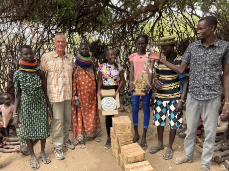 Das Foto zeigt 5 Vertreter des Dorfes Nakolonyoi mit Achim und Patricia; Patricia hält einen Karton mit Milchtüten in der Hand. Links eine junge Frau in einem grün-blau gemusterten Kleid mit farbigen Halsringen, daneben Achim der freundlich lächelt. Neben ihm Akatoroy Ngichwae, die Dorfsprecherin und Köchin, und dann Patricia, die einen offenen Karton mit runden, weißen Milchpackungen hält. Im Vordergrund stapeln sich mehrere braune Kartons mit der Aufschrift "FRESH MILK". Die Stimmung wirkt positiv und hoffnungsvoll, mit Lächeln und direkten Blicken in die Kamera.
