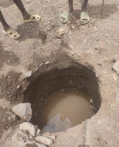 Das Foto zeigt die kläglichen Wasserreste in einem Loch im Dorf Nakolonyoi. Im Vordergrund sind zwei Paar Beine und Füße in einfachen Flip-Flops zu sehen, die am oberen Rand eines etwa meterbreiten, handgegrabenen Lochs im staubigen Boden stehen. Das Loch führt senkrecht nach unten und enthält am Grund eine kleine Pfütze trüben, schlammigen Wassers, umgeben von lockeren Erdbrocken und Steinen. Die Szene vermittelt Trockenheit und Armut in der ruralen, Turkana Region.