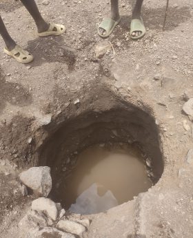 Das Foto zeigt die kläglichen Wasserreste in einem Loch im Dorf Nakolonyoi. Im Vordergrund sind zwei Paar Beine und Füße in einfachen Flip-Flops zu sehen, die am oberen Rand eines etwa meterbreiten, handgegrabenen Lochs im staubigen Boden stehen. Das Loch führt senkrecht nach unten und enthält am Grund eine kleine Pfütze trüben, schlammigen Wassers, umgeben von lockeren Erdbrocken und Steinen. Die Szene vermittelt Trockenheit und Armut in der ruralen, Turkana Region.