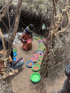Das Foto zeigt 1 Frau aus dem Dorf Nakolonyoi in ihrer einfachen Manyatta Küche, wie sie die Teller die wir BlackAndWhite gekauft hatten mit Mais und Bohnen befüllt. Die Frau kniet auf dem staubigen Boden einer traditionellen Hütte aus verflochtenen Zweigen und Ästen, trägt ein oranges Tuch mit farbigen Perlenhalsringen und konzentriert sich darauf, aus einem Topf (nicht sichtbar) portionsweise den Brei auf bunte Plastikteller in Pink, Blau und Grün zu verteilen. Vor ihr liegt eine gebogene Reihe von etwa 10-15 Tellern, einige bereits gefüllt mit gelblichen Körnern und Bohnen, was auf eine Mahlzeitenvorbereitung für die Gemeinschaft hinweist. Links und rechts sind Teile anderer Personen sichtbar, die sitzen, und die Szene ist in natürlichem Licht beleuchtet, das durch die offene Struktur der Hütte fällt. Die Atmosphäre vermittelt Rustikalität, harte Lebensbedingungen in einer arid afrikanischen Region, aber auch Dankbarkeit für die humanitäre Hilfe durch die gelieferten Utensilien und Lebensmittel.