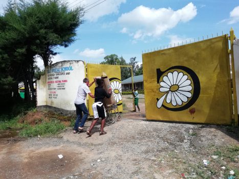 Achim und Sarah schieben einen gespendeten gebrauchten Rollstuhl mit einer Kartonbox die einen der brandneuen Rollstühle enthält durch das gelbe Tor der Daisy Special School für behinderte Kinder, das mit großen Gänseblümchen-Malereien verziert ist, unter einem sonnigen Himmel in einer ländlichen Umgebung.