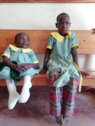 Zwei junge Kinder sitzen auf einer Holzbank, gekleidet in hellgrüne karierte Schuluniformen mit gelben Kragen. Eines lächelt verschmitzt, das andere schaut freuundlich neugierig nach vorne