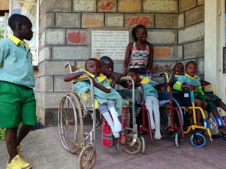 Patricia von BlackAndWhite steht lächelnd hinter einer Gruppe Kidsin hellgrünen Schuluniformen mit gelben Kragen, einige stehend und andere in den neuen Rollstühlen sitzend, posierend vor einer Steinmauer mit dem Schild „DAISY RESOURCE CENTRE FOR HANDICAPPED CHILDREN“ in einer Outdoor-Umgebung.