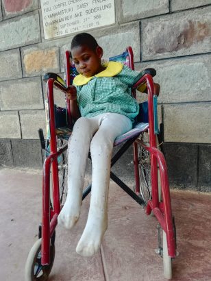 Ein junges Kid aus dem Daisy Center, im neuen roten Rollstuhl sitzend, gekleidet in eine hellgrüne karierte Schuluniform mit gelbem Kragen und weißen Strümpfen, und neugierig den Rollstuhl betrachtend