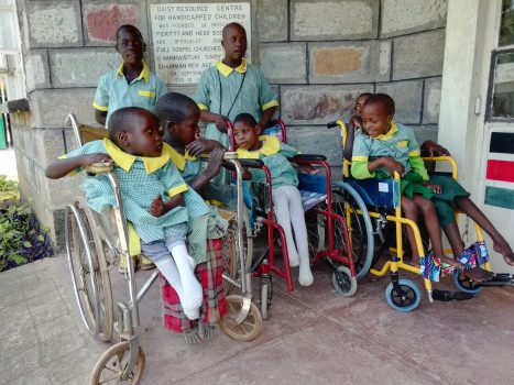 Ein paar der Kids in hellgrünen Schuluniformen mit gelben Kragen, einige stehend und andere teilen sich die brandneuen Rollstühle