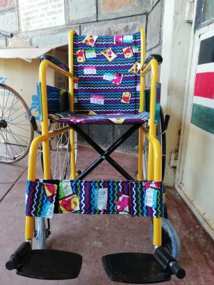 Ein bunter Kinderrollstuhl mit gelbem Rahmen steht vor einer gemauerten Wand. Sitz und Rückenlehne bestehen aus farbig gemustertem Stoff mit Tiermotiven. Der Rollstuhl ist neu und wirkt unbenutzt